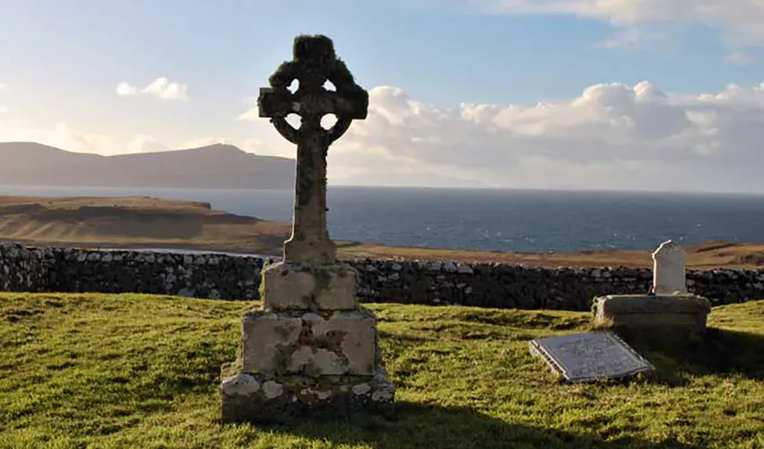 A stone Celtic Cross sits in graveyard with a view of mountains and the sea in the background