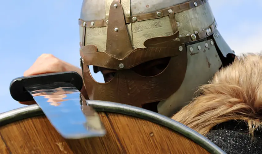 Dave dressed as Macbeth with a close up of his helmeted face, piercing eyes through the visor holding a sword on top of a shield