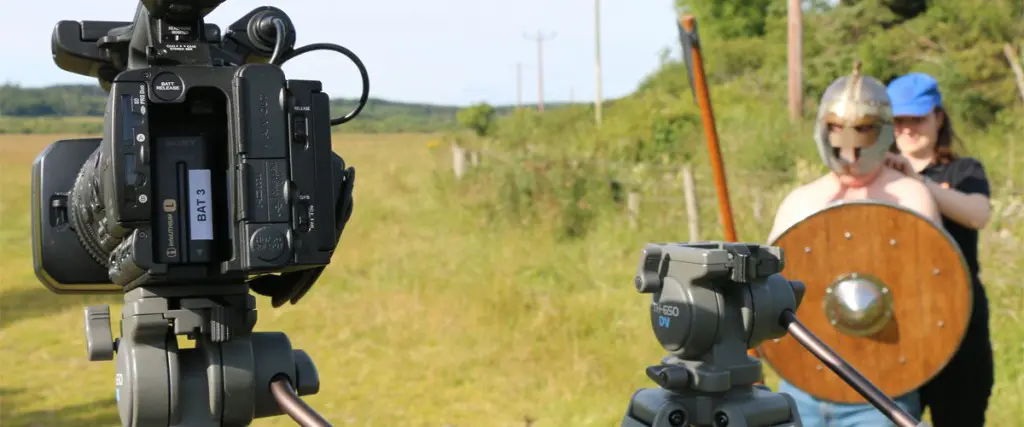 A camera on the left is off and pointingat afield with Sara and Paul on the right getting ready for a photo shoot