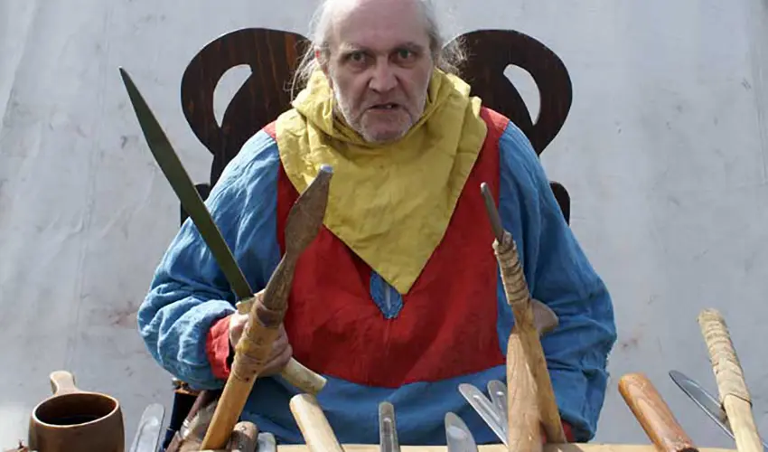 An aged man brandishes a short sword in front a table of various replica weapons of the period